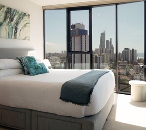 A bedroom with a large bed, a view of a cityscape through floor-to-ceiling windows, and a city skyline visible outside. - L18 Stunning 3 BRM Apt with Beach Views (Broadbeach)