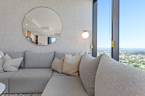 Ein modernes Wohnzimmer mit einer großen Eckcouch, einem runden Spiegel und Blick auf eine Stadtlandschaft durch große Fenster.