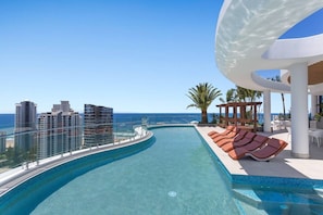 A pool with a curved edge, lounge chairs, and a view of the ocean and buildings. - Stunning L19 Dlx 3 Bedroom Apt w/ Hinterland Views (Broadbeach)