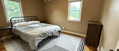 A bedroom with a bed, a ceiling fan, two windows, a radiator, and a door.