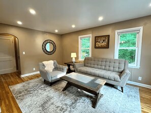 Ein Wohnzimmer mit grauem Sofa, einem grauen Sessel, einem Couchtisch, einem runden Spiegel und zwei Fenstern mit Blick auf Grünflächen.