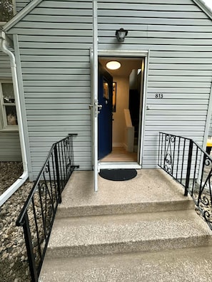 A set of concrete steps leading to a door with a number '813' on the wall, a small outdoor area with a black mat, and a metal railing on the left. - Kutzky Duplex: Walk to Mayo 2BR with Free Parking (Rochester)