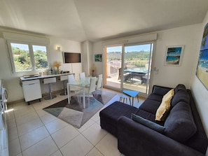 A modern living room with a sofa, a dining table with chairs, a kitchen area, and large windows offering a view of the landscape. - 2 bedrooms, sea view, air conditioning,all on foot (ST MANDRIER SUR MER)