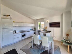 A modern kitchen with white cabinetry, a built-in oven, and a dining area with a glass table and white chairs. - 2 bedrooms, sea view, air conditioning,all on foot (ST MANDRIER SUR MER)
