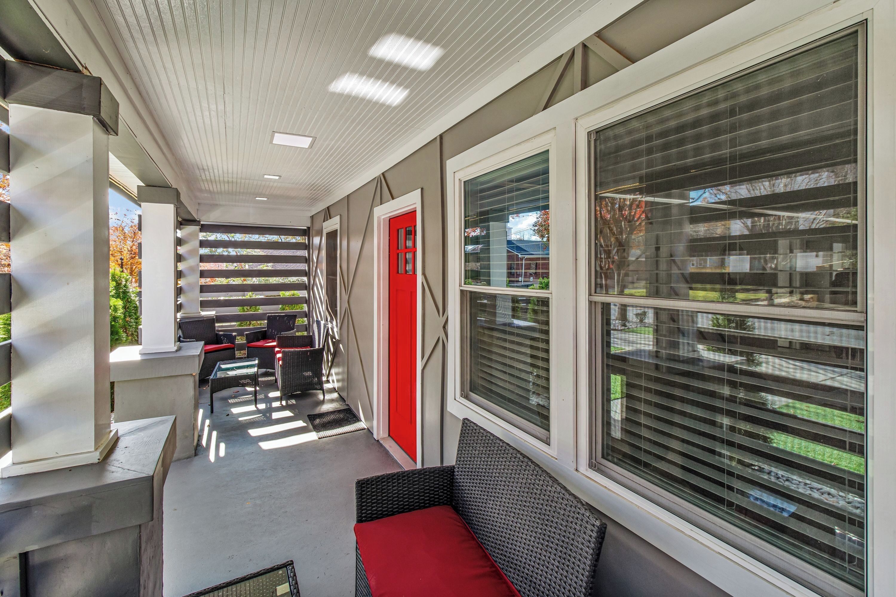 A covered outdoor seating area with chairs and tables, a red door, and large windows with blinds.