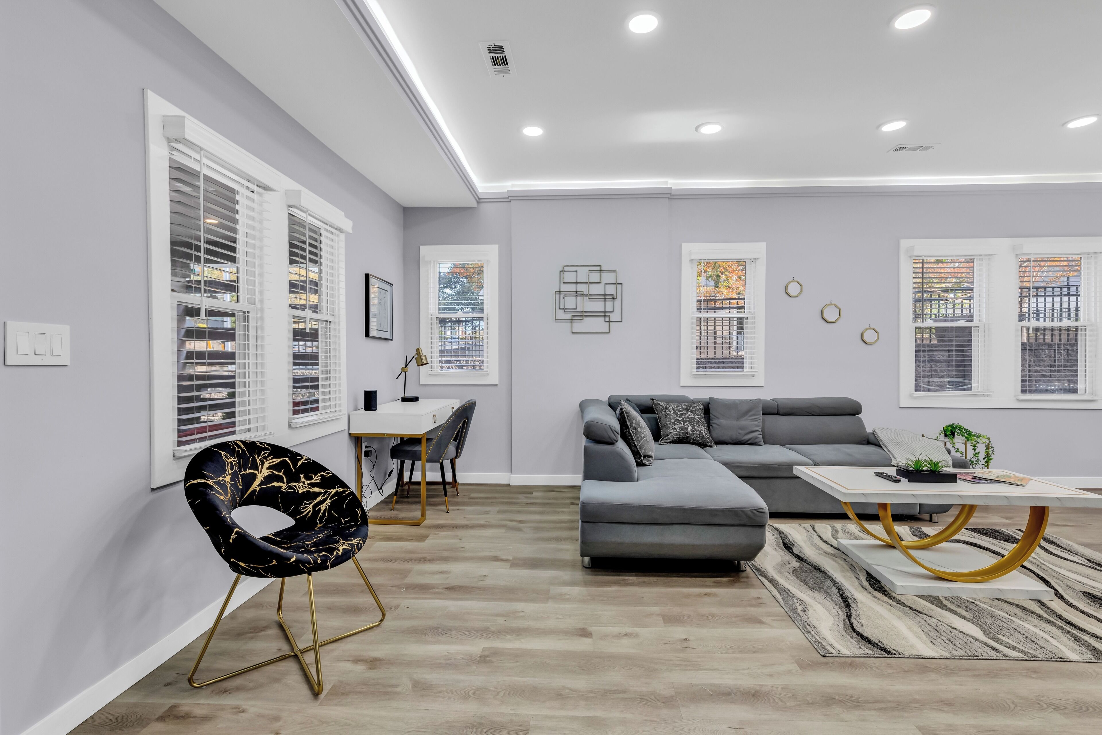 A modern living room with a grey sofa, a dining area with a white table, and a black chair with gold legs.