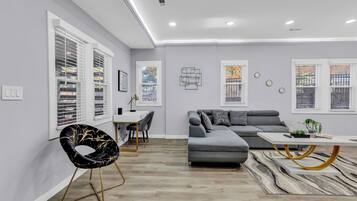 A modern living room with a grey sofa, a dining area with a white table, and a black chair with gold legs.