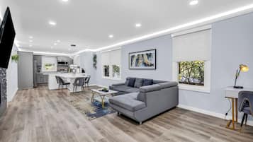 A modern living room with a grey sofa, a dining area, and a kitchen in the background.