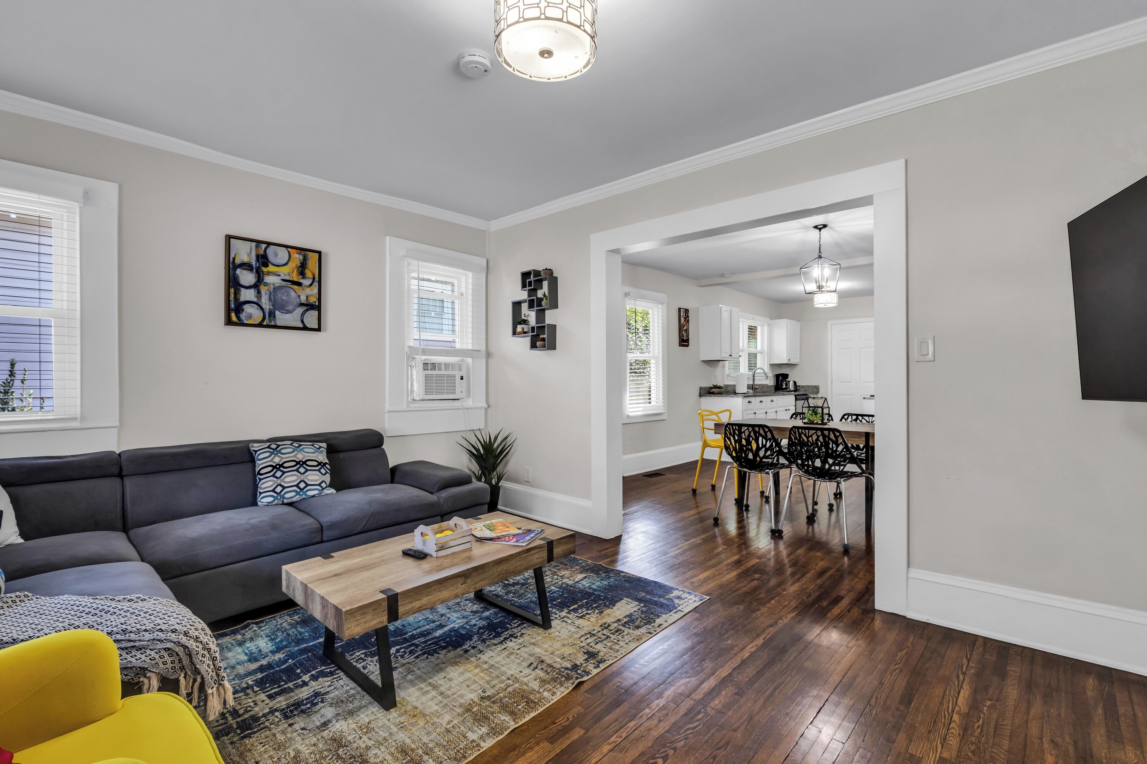 A modern living room with a gray sofa, a coffee table, and a rug. There is a kitchen area with a dining table and chairs visible through an open doorway.