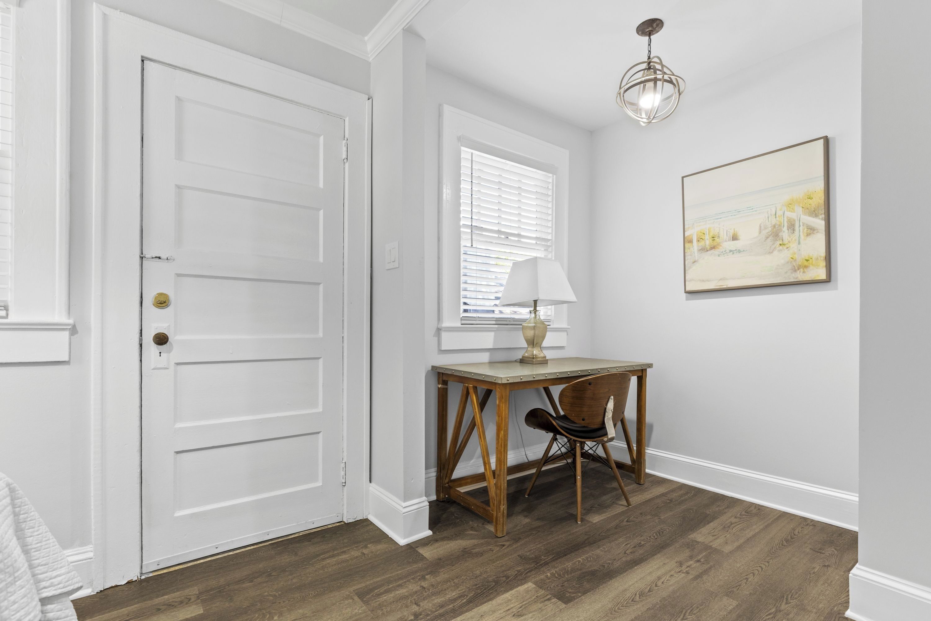 A room with a white door, a desk with a lamp, a chair, and a painting on the wall.
