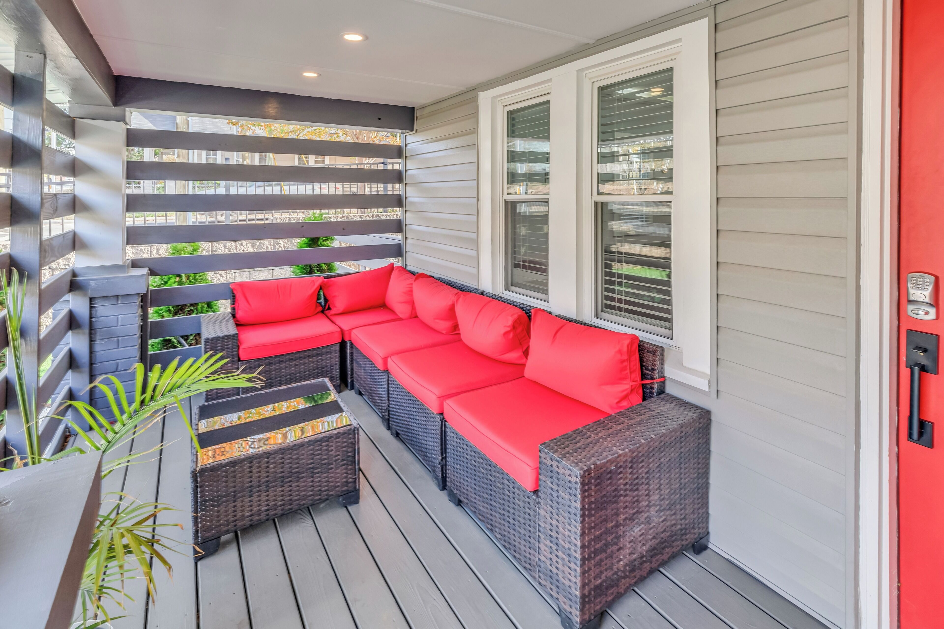 A covered outdoor seating area with a sectional sofa and a small table, surrounded by a wooden deck and potted plants.