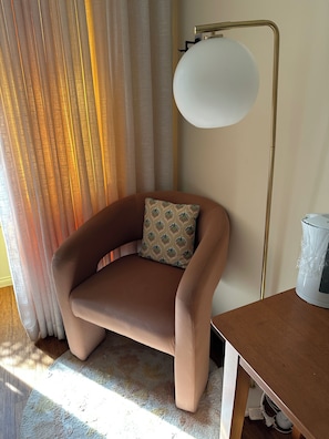 Una habitación con un sillón marrón, un cojín estampado, una lámpara de pie y una mesa.
