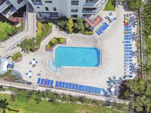Eine Vogelperspektive auf ein Hotel mit einem rechteckigen Swimmingpool, umgeben von blauen Liegestühlen und einem überdachten Sitzbereich.