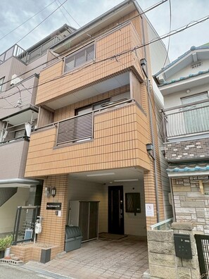 A multi-story residential building with a tiled facade, a visible entrance, and a small outdoor area with a trash bin. - Neo Universal 04 (Osaka, Konohana, Nishikujo)