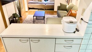 A compact kitchen with a built-in microwave, a small table, and a sofa in the background. - Neo Universal 04 (Osaka, Konohana, Nishikujo)