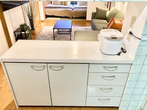 A compact kitchen with a built-in microwave, a small table, and a sofa in the background. - Neo Universal 04 (Osaka, Konohana, Nishikujo)