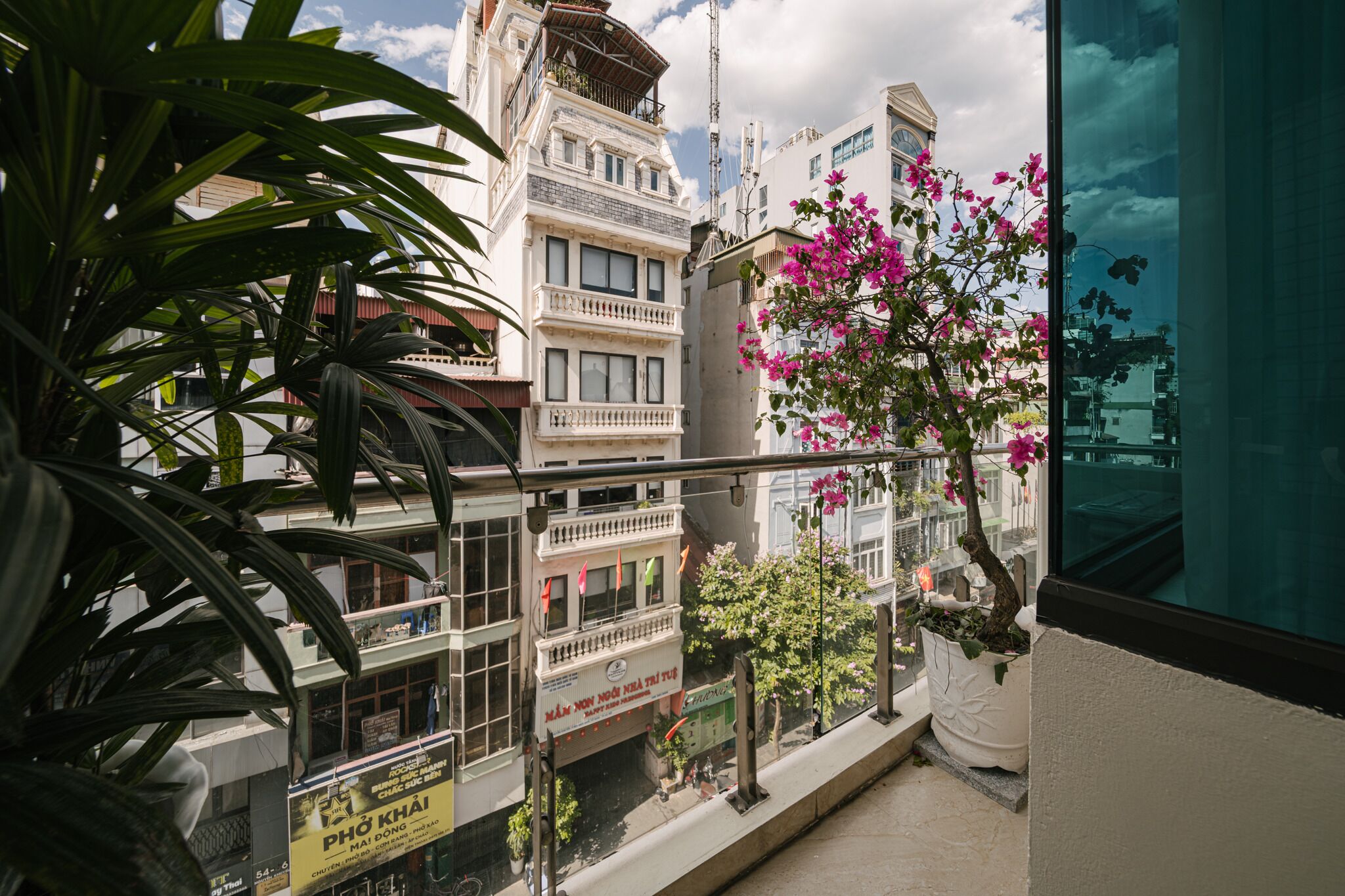 A balcony with a view of a building with multiple floors, a potted plant with pink flowers, and a sign with text.