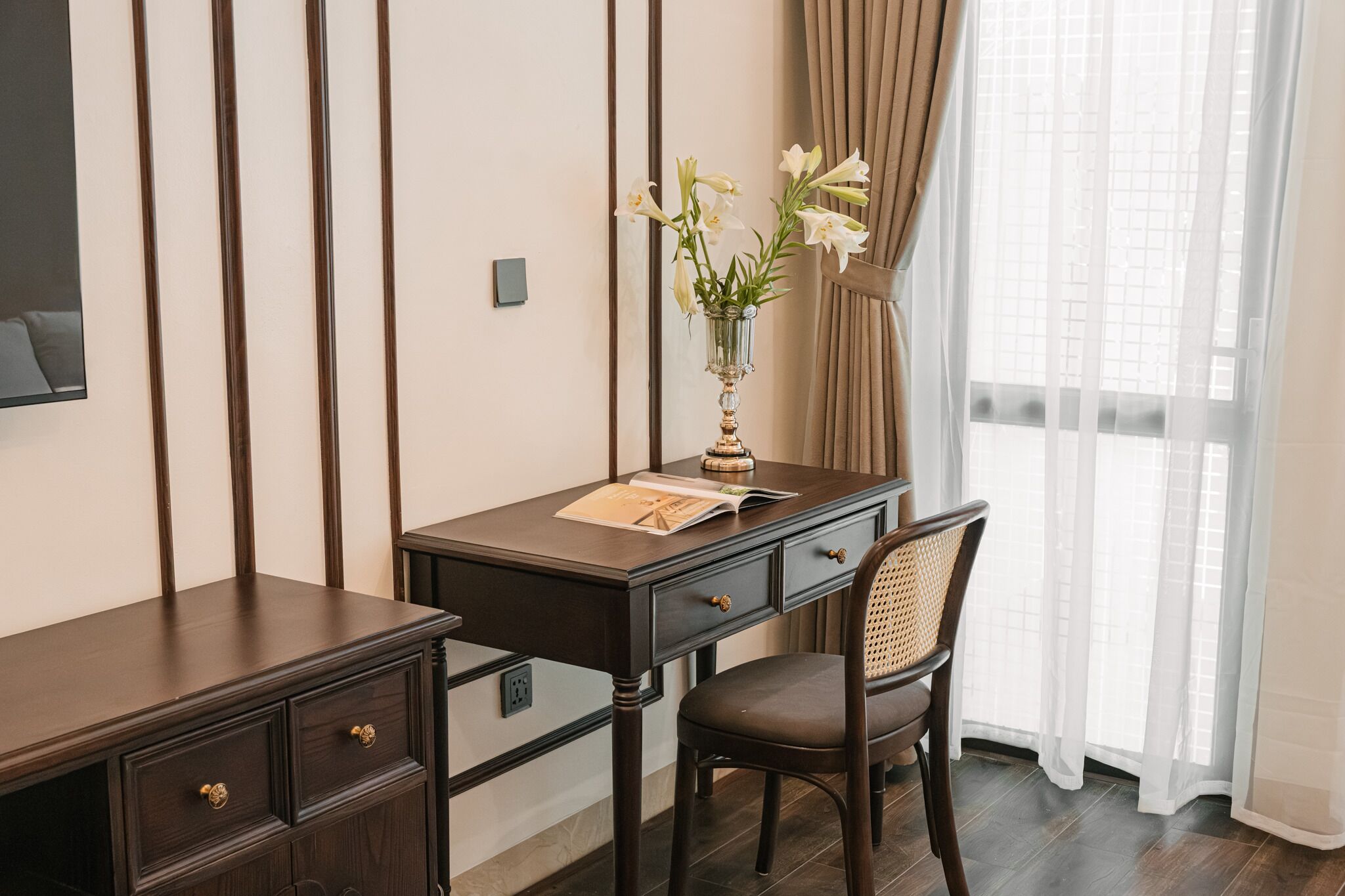 Uma mesa de madeira escura com um vaso de flores brancas, uma cadeira e uma janela com cortinas translúcidas.