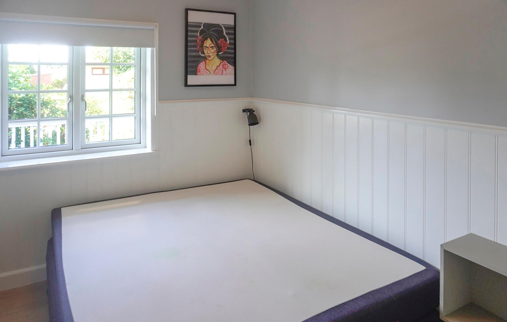 A bedroom with a single bed, a window, a wall-mounted lamp, and a framed picture.
