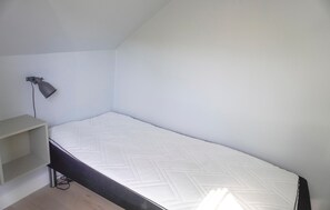 Ein kleines Zimmer mit einem Einzelbett, einer schlichten weißen Wand, einem Schreibtisch mit Lampe und einem schlichten Boden.