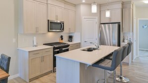 A modern kitchen with light wood cabinets, stainless steel appliances, a central island with bar stools, and pendant lighting. - 4BR Cozy Home in  Calgary with Hot Tub (Calgary)