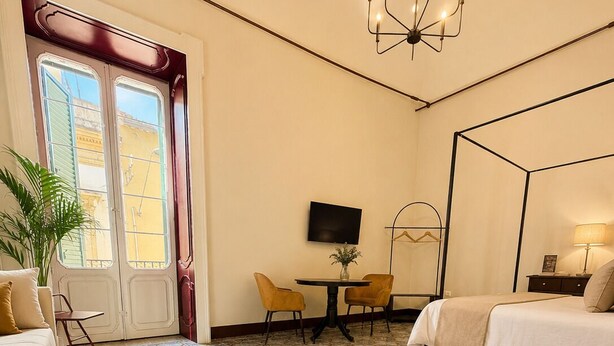 A hotel room with a four-poster bed, a TV on the wall, a dining table with chairs, and a large window with shutters.