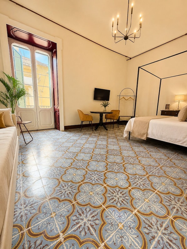 A hotel room with a four-poster bed, a TV on the wall, a dining table with chairs, and a large window with shutters.