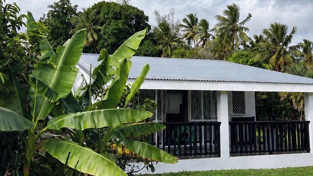 A white building with a black railing, surrounded by lush greenery and palm trees. - ‘Ūtū Retreat 
 a peaceful open-plan studio designed for rest (Avarua)