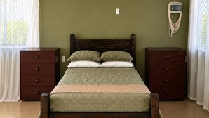 A bedroom with a wooden bed, two nightstands, a wall-mounted heater, and a window with curtains. - ‘Ūtū Retreat 
 a peaceful open-plan studio designed for rest (Avarua)
