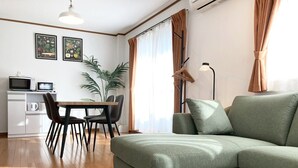 A modern living room with a green sofa, a dining table with chairs, a kitchen area with a microwave, and a large plant. - Neo Universal 05 (Osaka, Konohana, Nishikujo)