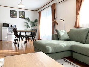 Ein modernes Wohnzimmer mit einem grünen Sofa, einem Esstisch mit Stühlen, einer Küchenzeile mit einer Mikrowelle und einer großen Pflanze.