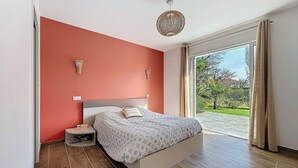 Ein Schlafzimmer mit einem Bett, einem Nachttisch, einer Wandlampe und Blick auf einen Garten durch eine Schiebetür.