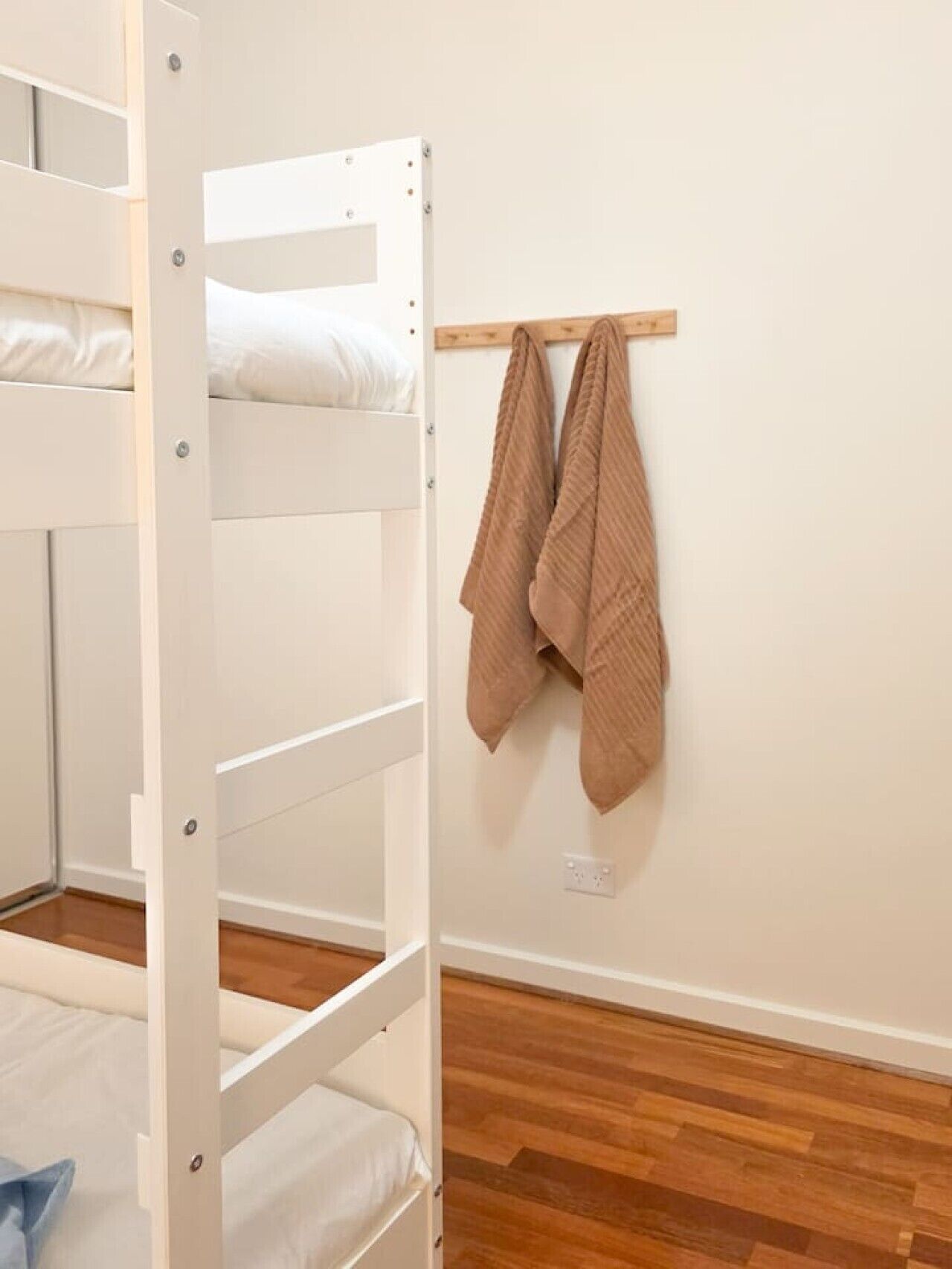 A bunk bed with a wooden frame and white bedding, a wooden shelf with a brown towel, and a wooden floor.
