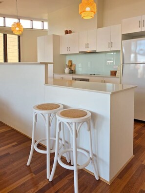 A modern kitchen with white cabinetry, a central island, two bar stools with woven seats, and pendant lighting.