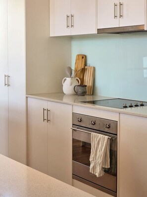 Eine Küche mit weißen Schränken, einem Edelstahlbackofen und einer Arbeitsplatte mit Schneidebrett, Wasserkocher und Löffel.