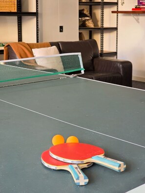 A ping pong table with two paddles and three balls in a room with a brown leather couch and metal shelving.