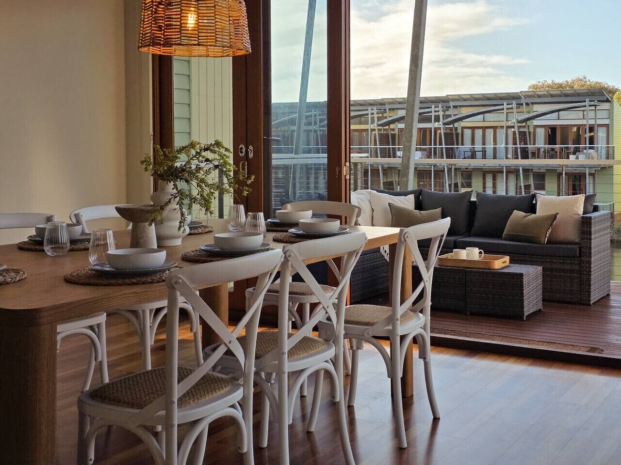 A dining area with a wooden table set for four, surrounded by white wicker chairs. A sofa and a small table with a tray are visible in the background.