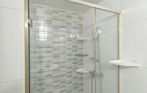 Una ducha con mampara de vidrio, pared con azulejos, lavamanos blanco y repisa de cristal.