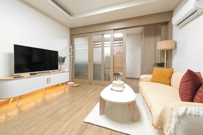 Una sala de estar moderna con un televisor de pantalla plana, una mesa de centro redonda, un sofá con cojines, una lámpara de pie y un ventanal corredero que da a un balcón.