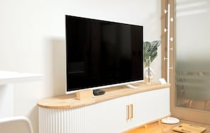 Une chambre moderne avec un téléviseur à écran plat fixé à un mur blanc, une console en bois dotée de poignées dorées et une plante en pot.