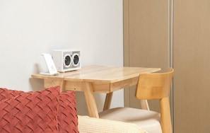 Une table en bois, sur laquelle se trouvent un haut-parleur blanc et un appareil blanc, un coussin rouge tissé et une chaise en bois.
