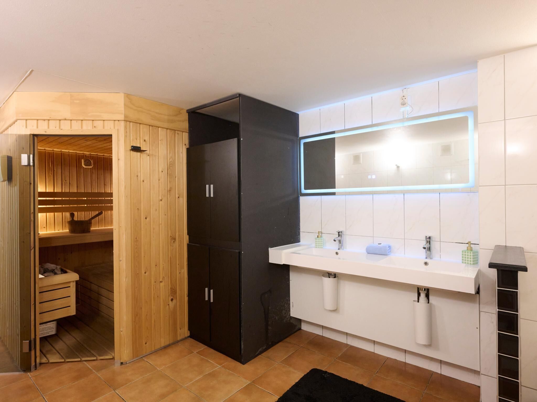 A modern bathroom with a wooden sauna, a black cabinet, a white sink with a mirror, and a black rug.