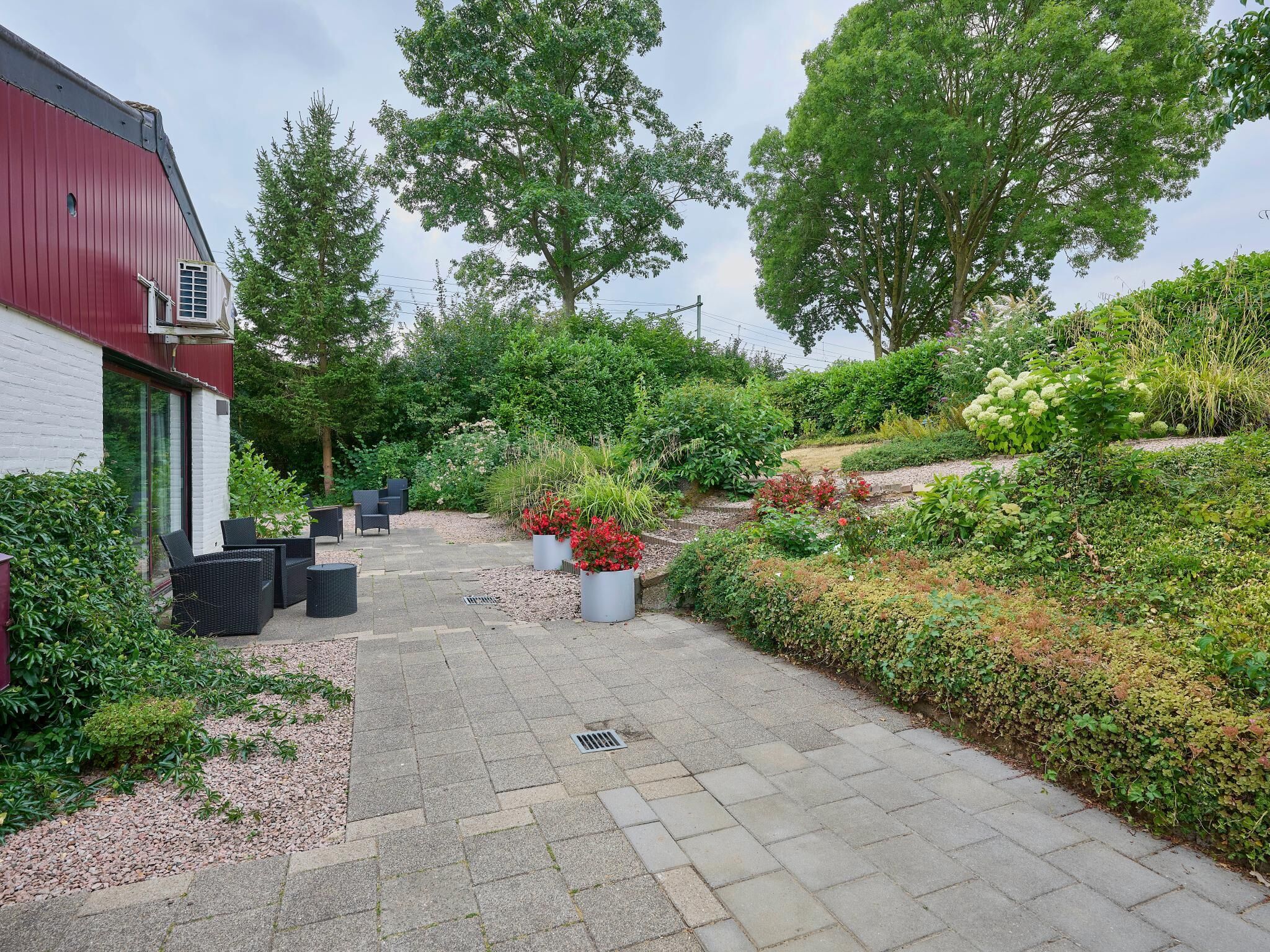 Een geplaveid pad dat leidt naar een gebouw met een rood dak, omgeven door weelderige begroeiing en verschillende planten.
