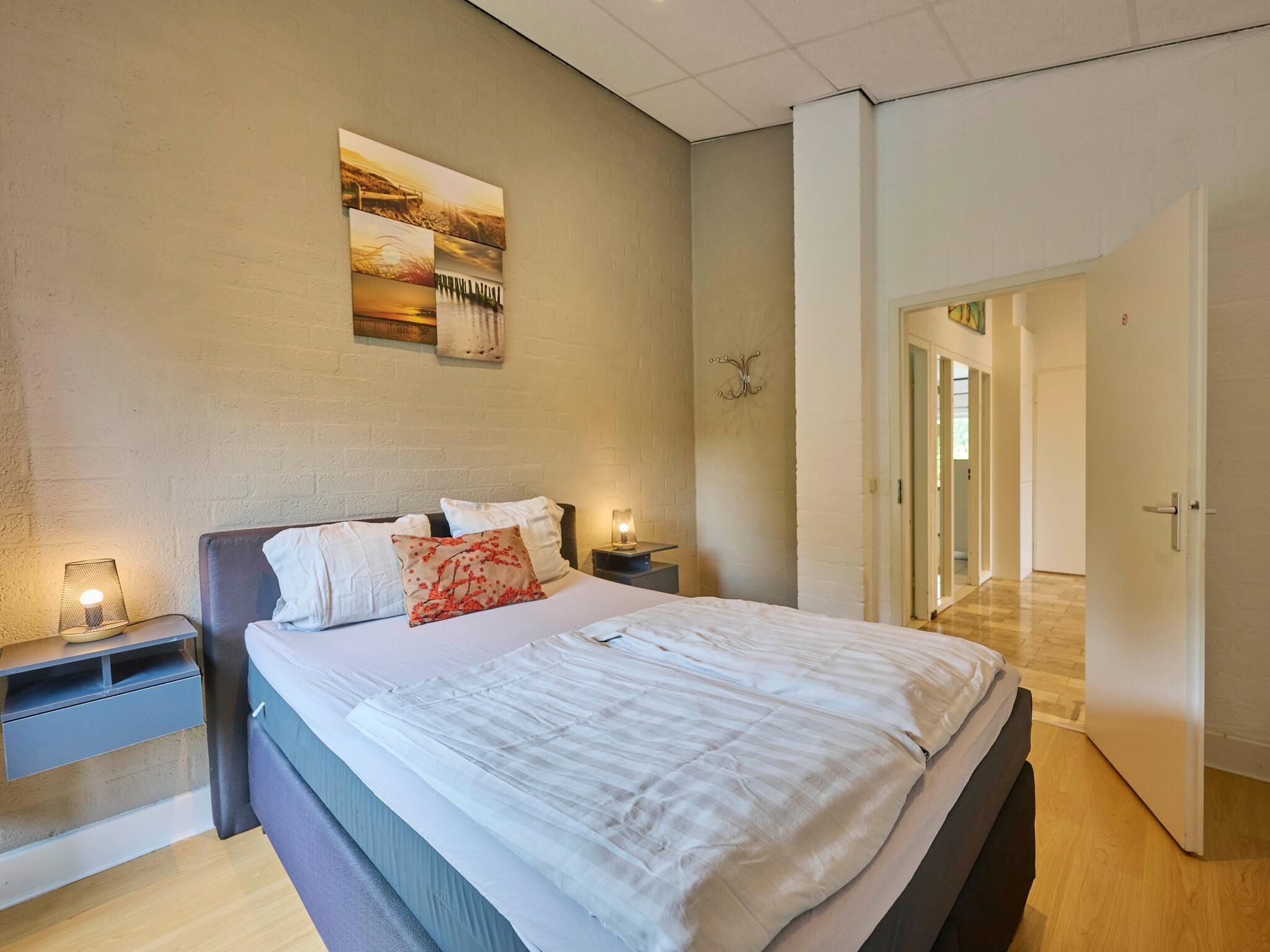 A bedroom with a bed, nightstands, a wall-mounted lamp, and a door leading to another room.