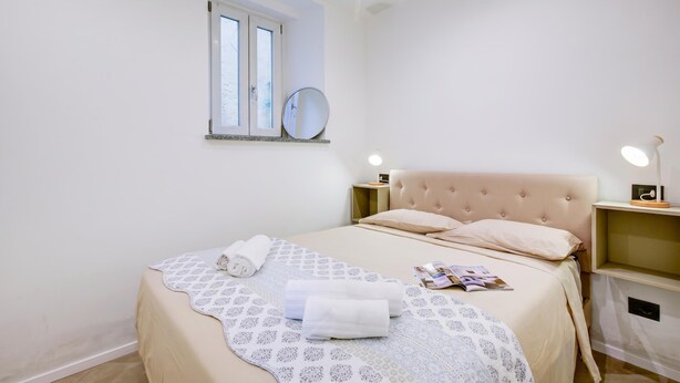 A bedroom with a bed, a window, a bedside table with a lamp, and a wall-mounted mirror. - La Porte Rouge 100 Mt From Lake Maggiore (Meina)