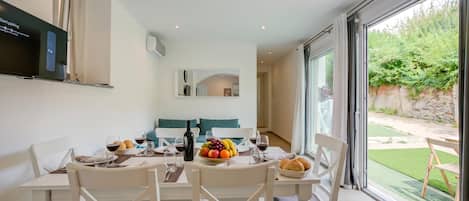 Un comedor con una mesa preparada para cuatro personas, un ventanal con vistas a un jardín y un reloj colgado en la pared.