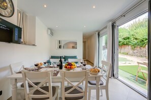 A dining area with a table set for four, a large window with a view of a garden, and a wall-mounted clock. - La Porte Rouge 100 Mt From Lake Maggiore (Meina)