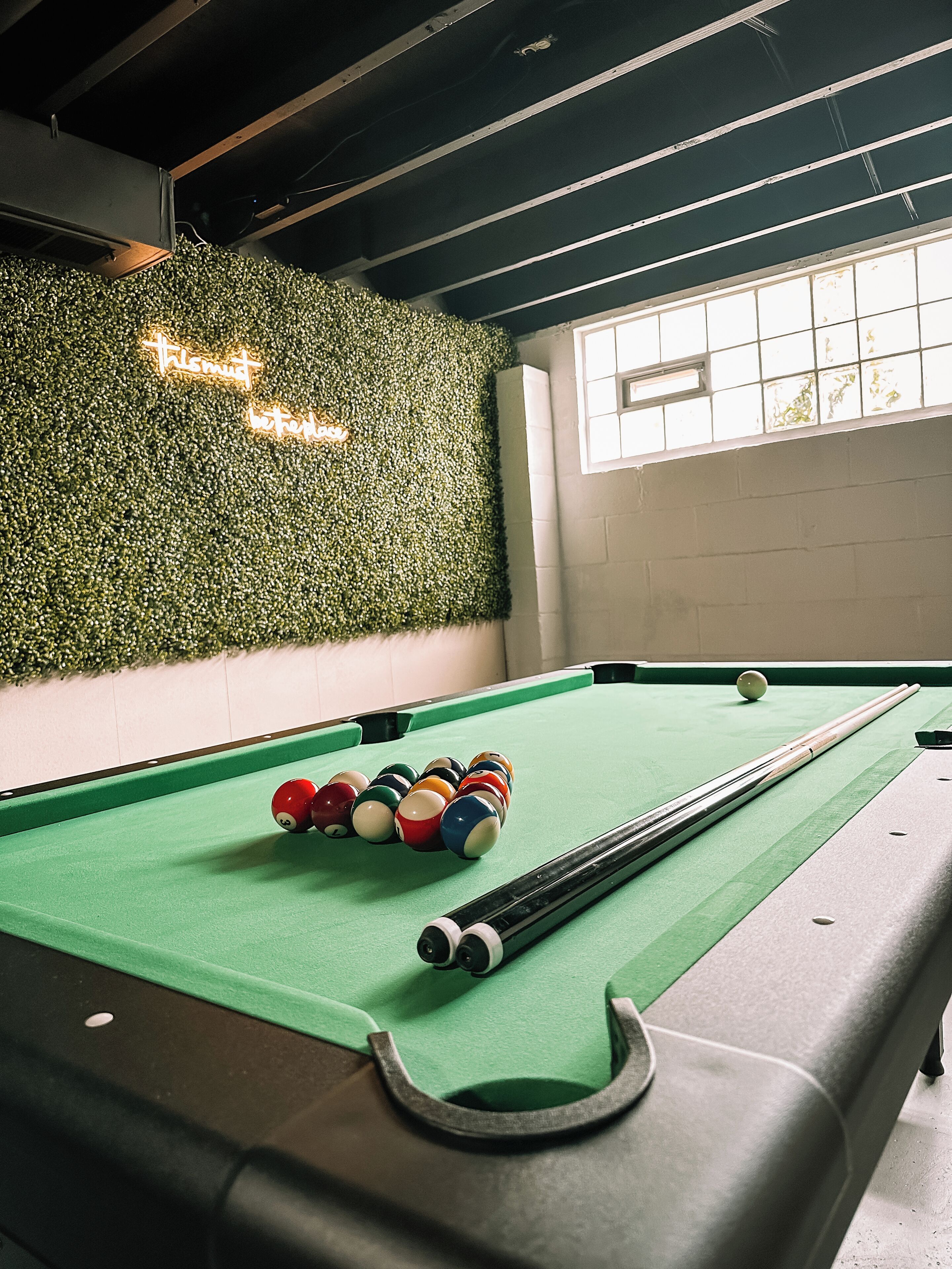 A pool table with balls and a cue stick, a green artificial grass wall, and a window with daylight.