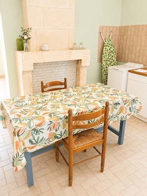 Ein Essbereich mit einem Tisch, bedeckt mit einer blumigen Tischdecke, zwei Holzstühlen, einem Kamin aus Ziegelsteinen und einer Kleiderstange.