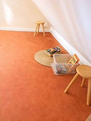 Ein Zimmer mit Teppichboden, einem kleinen Holzhocker, einer gewebten Matte und einer Plastikaufbewahrungsbox voller Bücher.